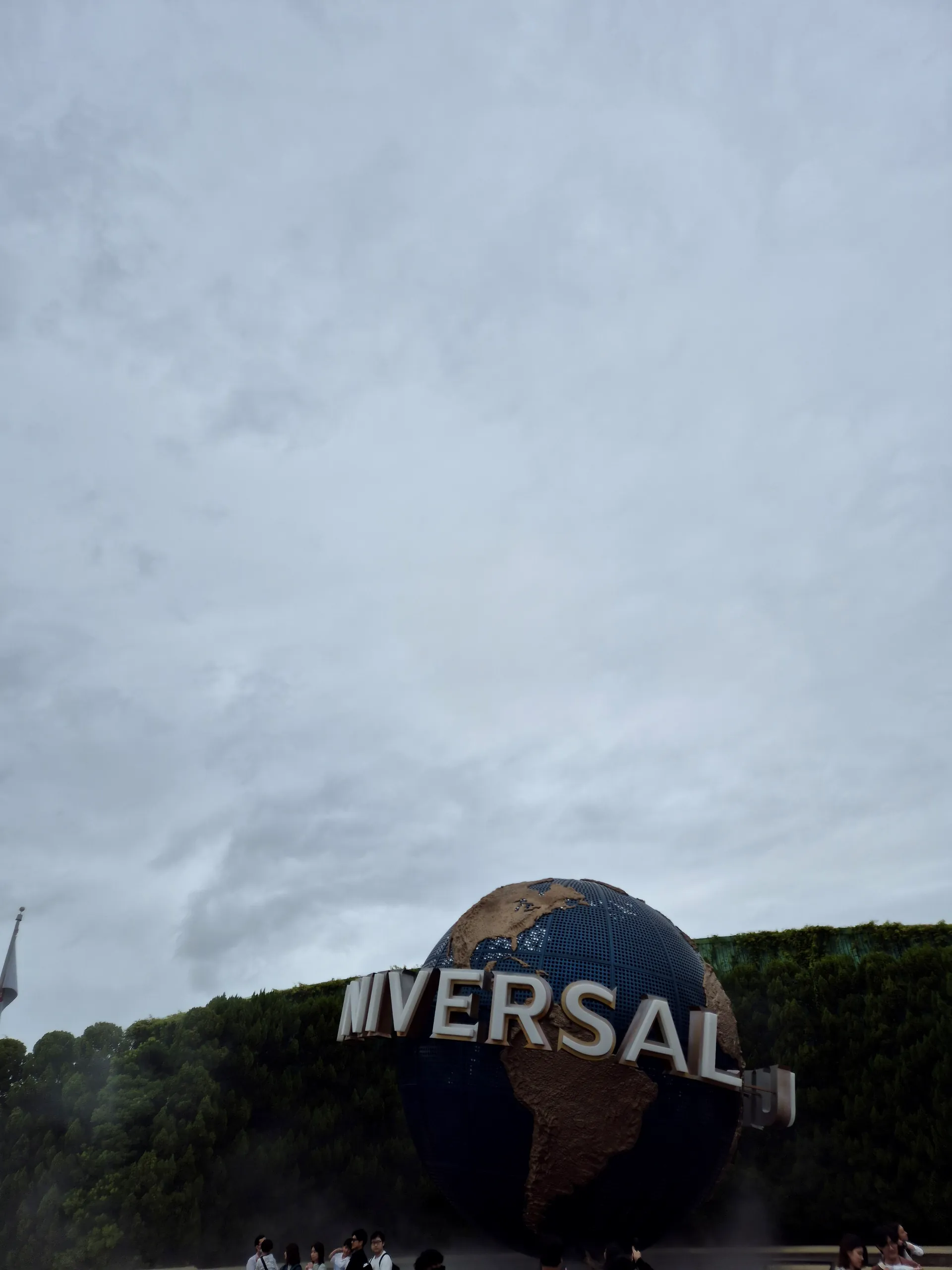 UNIVERSAL 글자가 둘러진 커다란 지구본과 사진 위쪽 대부분을 채운 잿빛 구름 하늘, 아래쪽의 초록 덤불