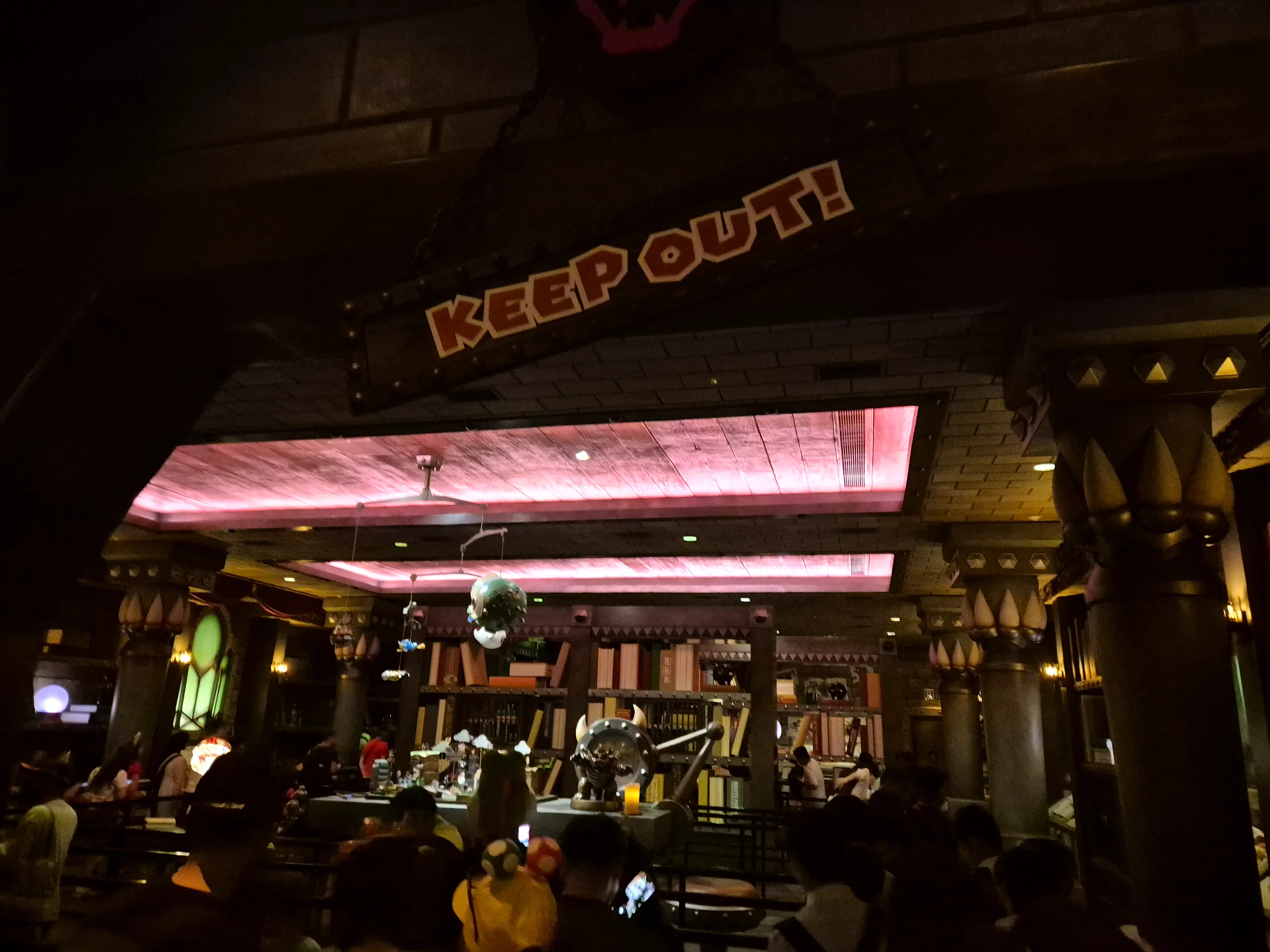 분홍빛 조명이 비치는 어두운 실내에 KEEP OUT! 간판과 책장, 소품이 있고 줄 선 사람들이 실루엣으로 보이는 쿠파 성 대기 공간